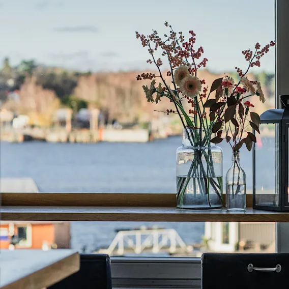 En glasvas med blommor och en svart lykta står på en fönsterbräda med utsikt över lugnt vatten och suddiga träd i bakgrunden.