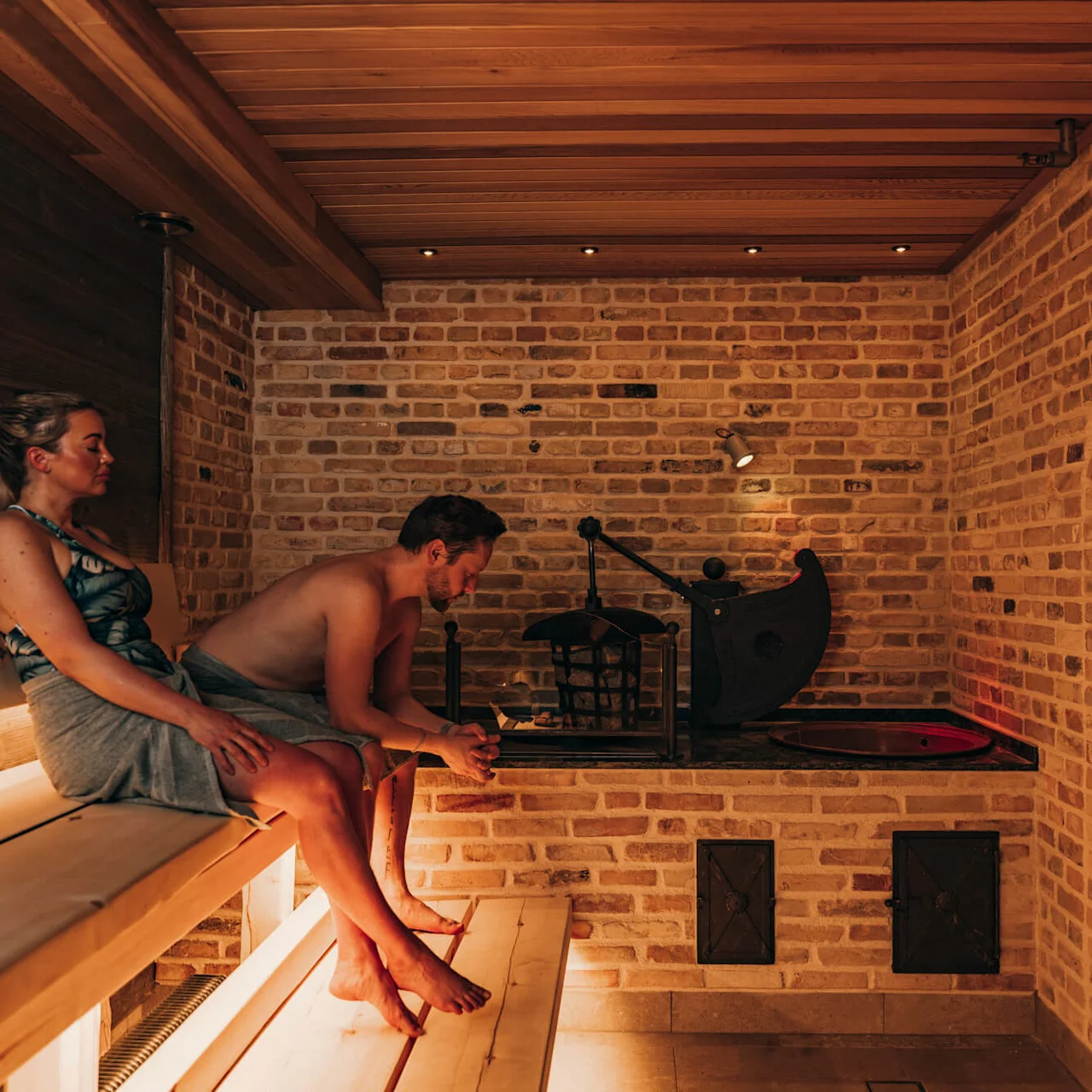Two people sit on a wooden bench in a sauna, relaxing against a backdrop of warm-toned brick walls. Dim lighting creates a cozy atmosphere.