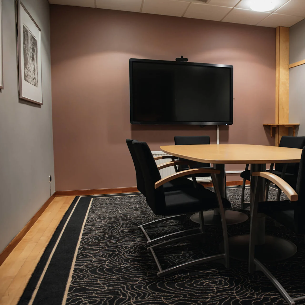 A conference room features a large monitor on a wall, a wooden table surrounded by black chairs, and framed art. The room has wooden floors and a patterned carpet.