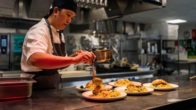 Chef plating dishes with a sauce in a professional kitchen, surrounded by stainless steel equipment and cooking utensils.