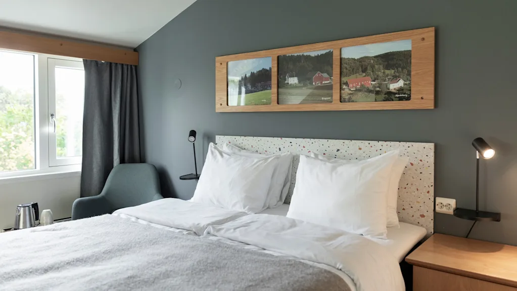 A cozy hotel room with a comfortable bed, framed landscape art, and a window offering a view of nature.