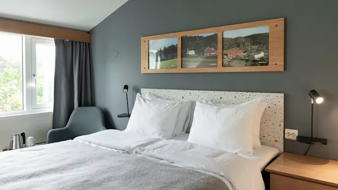 A cozy hotel room with a comfortable bed, framed landscape art, and a window offering a view of nature.