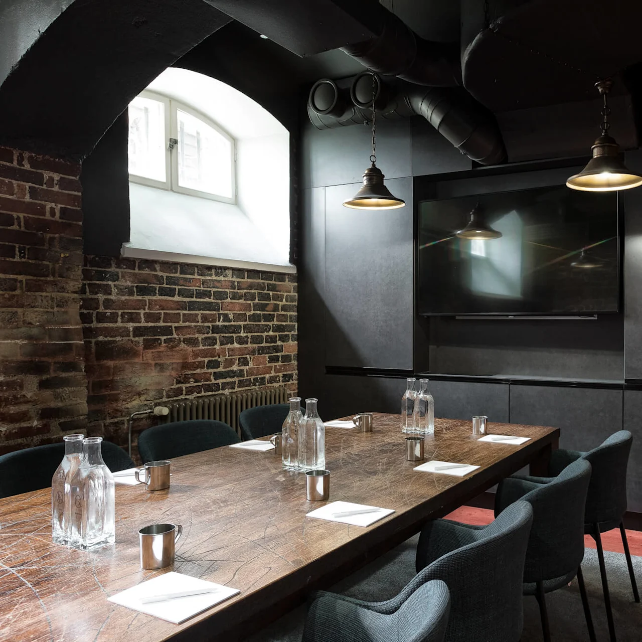 A wooden conference table with chairs, exposed brick walls, pendant lights, and a mounted screen against a dark backdrop.