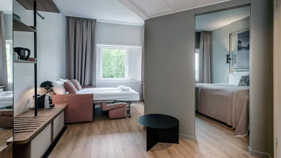 Et moderne hotellrom har en pent oppredd seng, lite salongbord og espressomaskin. Tregulv og stort vindu skaper en lys, innbydende atmosfære.