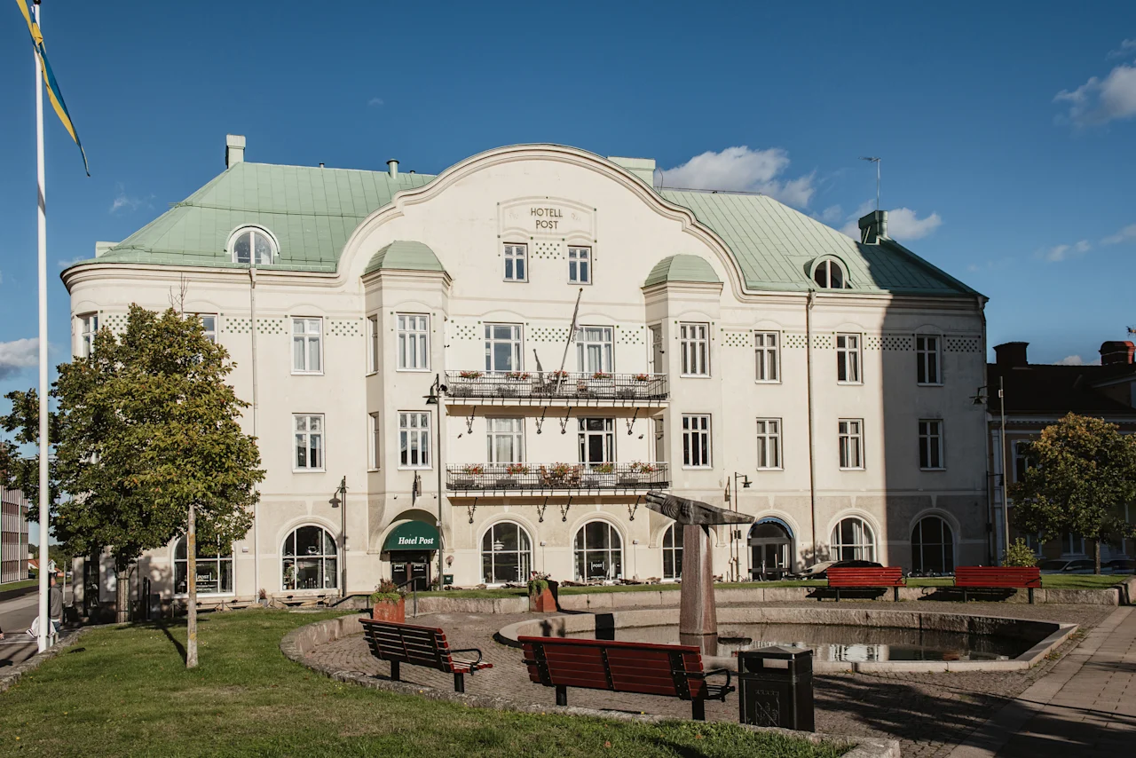 A grand white building with a green roof and balconies, labeled HOTELL POST and Hotel Post, stands behind a park with benches and a pond.