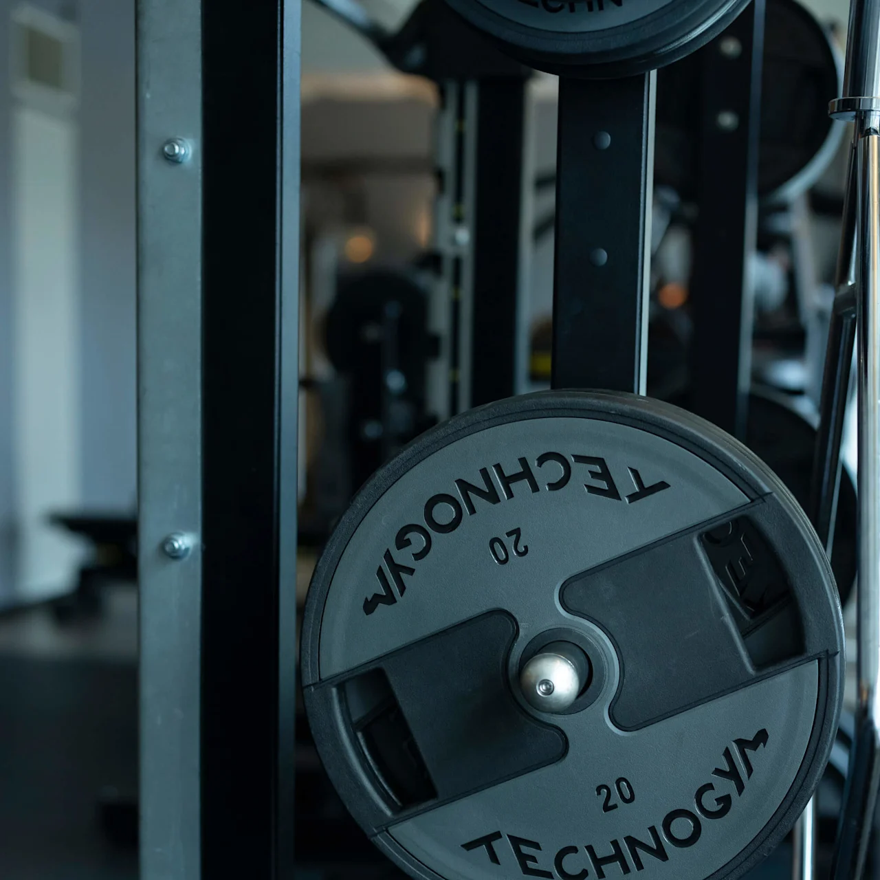 Painolevy lepää kuntosalilaitteessa, merkitty "TECHNOGYM 20". Ympäröivä alue näyttää muuta kuntosalilaitteistoa himmeästi valaistussa liikuntakeskuksessa.