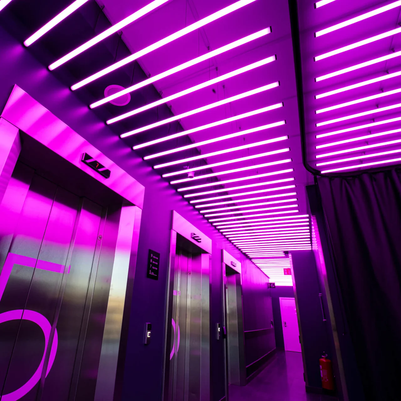 Elevatorer med lilla belysning oplyser gangen. Tal på elevatordøre viser "5". En moderne, levende atmosfære skabes af vandrette neonlys ovenfor.