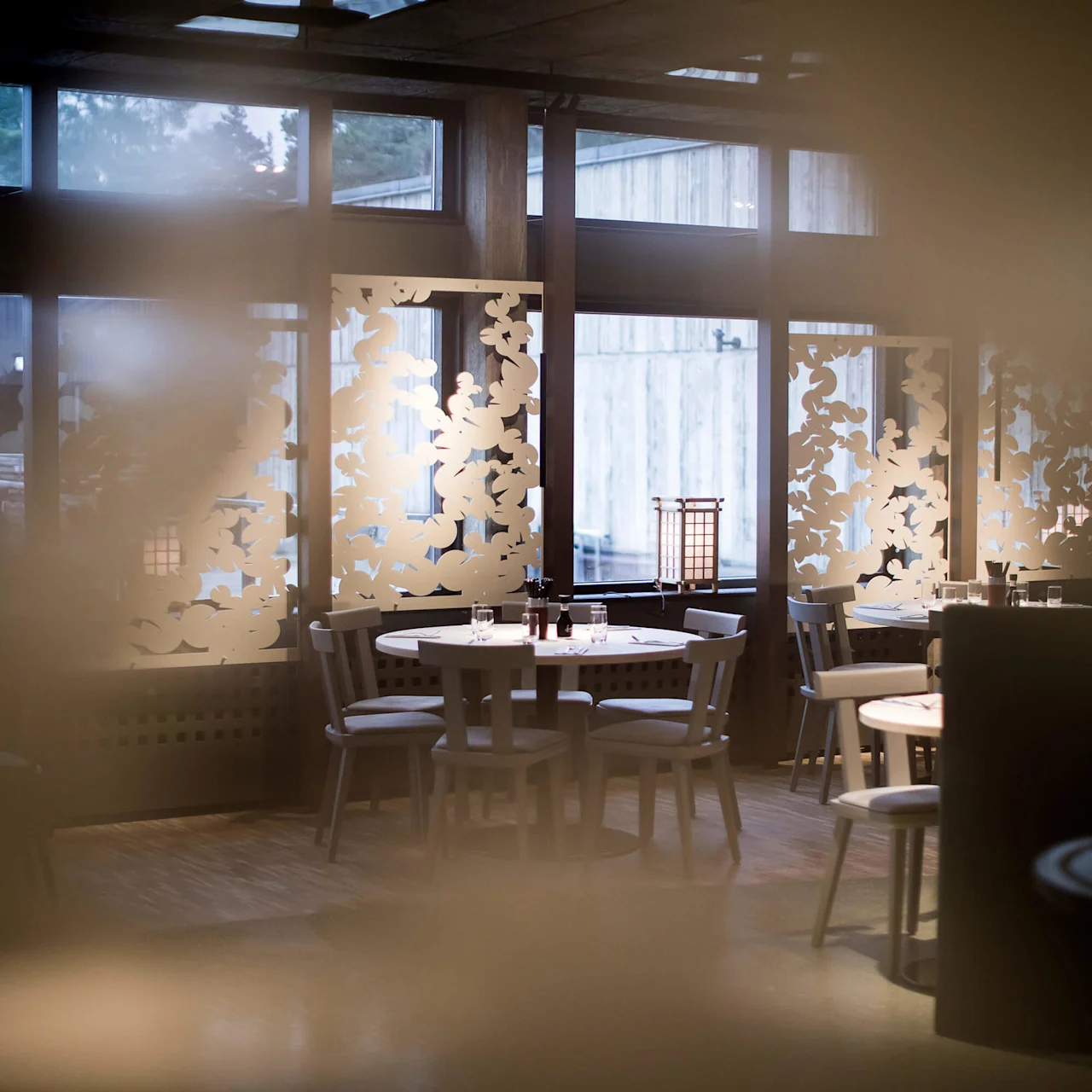 En dempet belyst restaurant med tomme bord, mønstrede skiller og store vinduer.