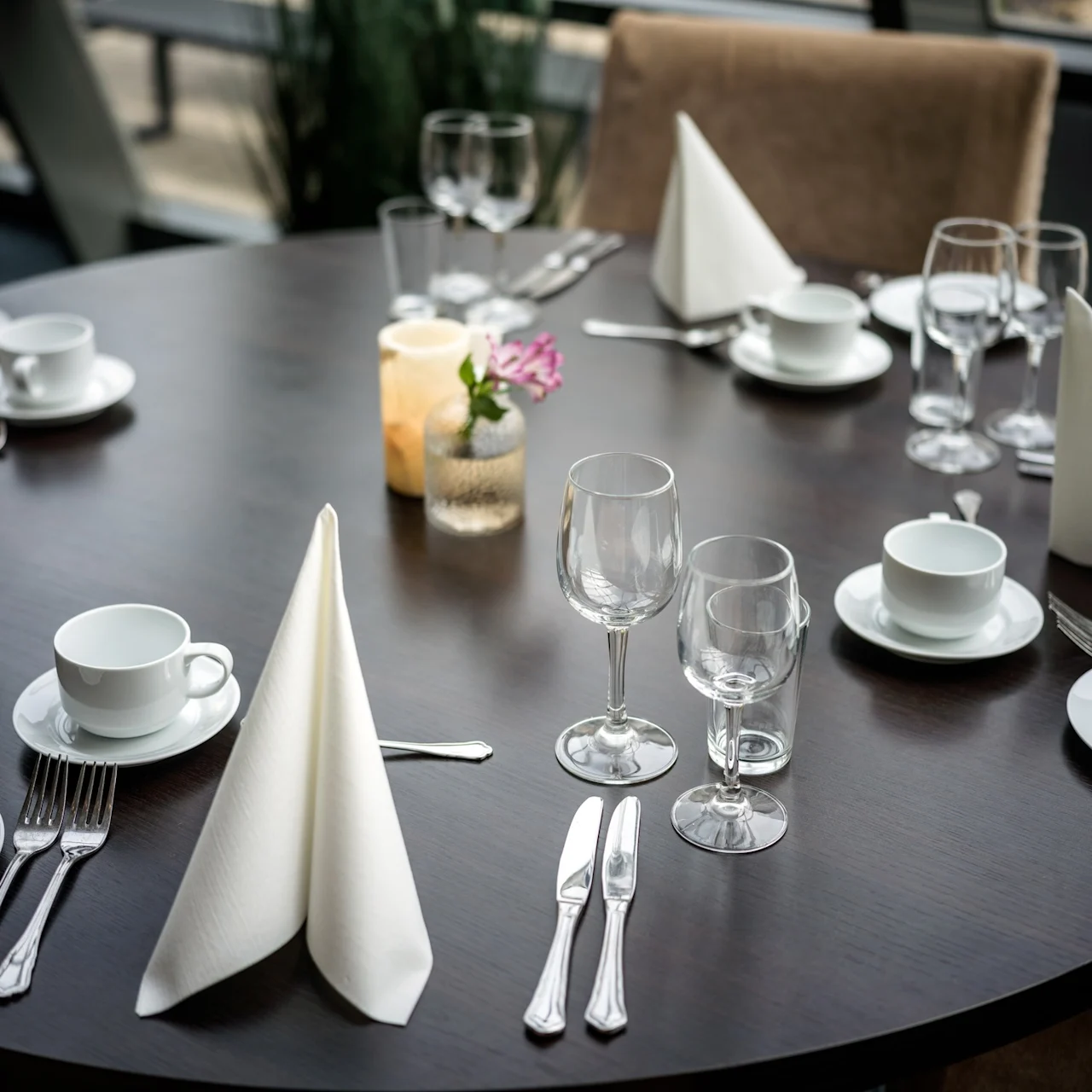 Runt matbord dukat för måltid; tallrikar, koppar, glas och servetter arrangerade, ljus och blomstervas i mitten. Ljus, modern interiör med naturligt ljus genom stora fönster.