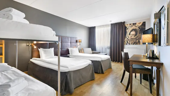 To pænt redte senge står i et lyst hotelværelse med et zebrabillede, gardiner, skrivebord, stol og vægmonteret TV. En køjeseng er i forgrunden.