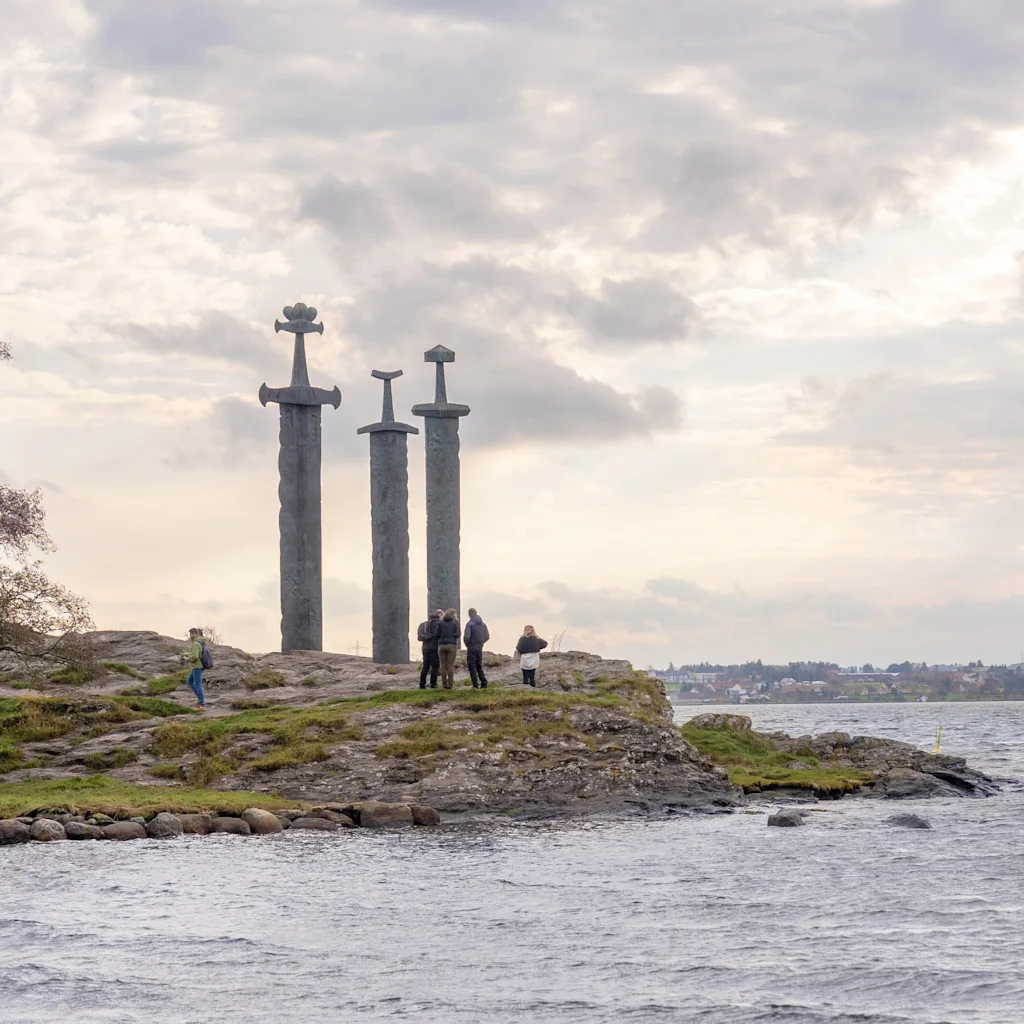 Kolme monumentaalista miekkaveistosta seisoo kallioisella rannalla, josta avautuu näkymä vesistöön pilvisen taivaan alla. Ihmiset tarkkailevat näkymää.