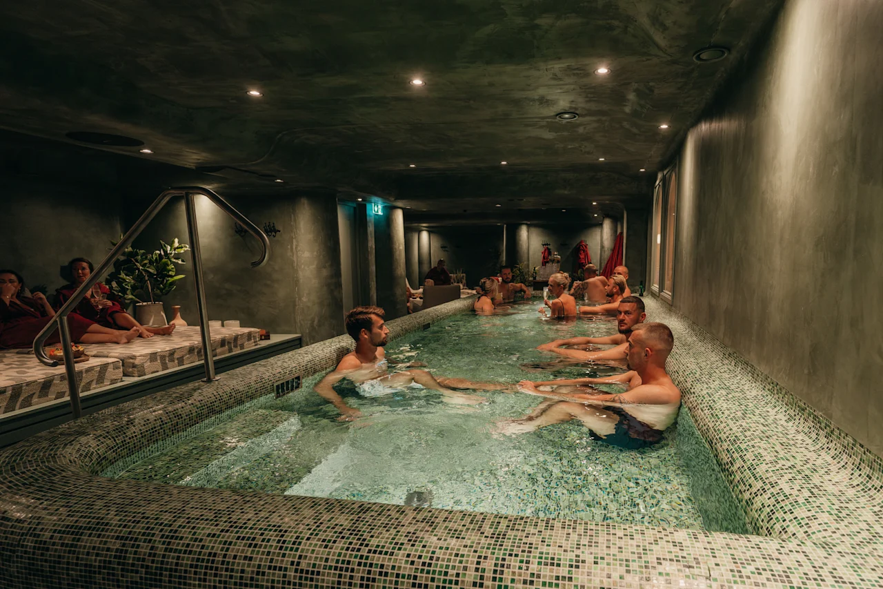 Vieraat rentoutuvat rauhallisessa, hämärästi valaistussa kylpyläaltaassa mosaiikkilaatoilla. Jotkut uivat vedessä, kun taas toiset rentoutuvat altaan äärellä olevilla lepotuoleilla.