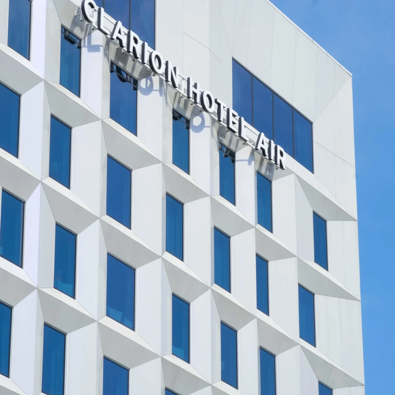En moderne hvit bygningsfasade med geometriske vinduer, som viser teksten "CLARION HOTEL AIR" mot en klar blå himmel.