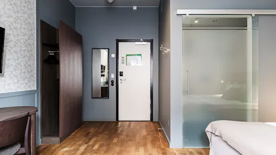 Ett hotellrum med en säng, en duschplats med glasväggar och en utgångsdörr.