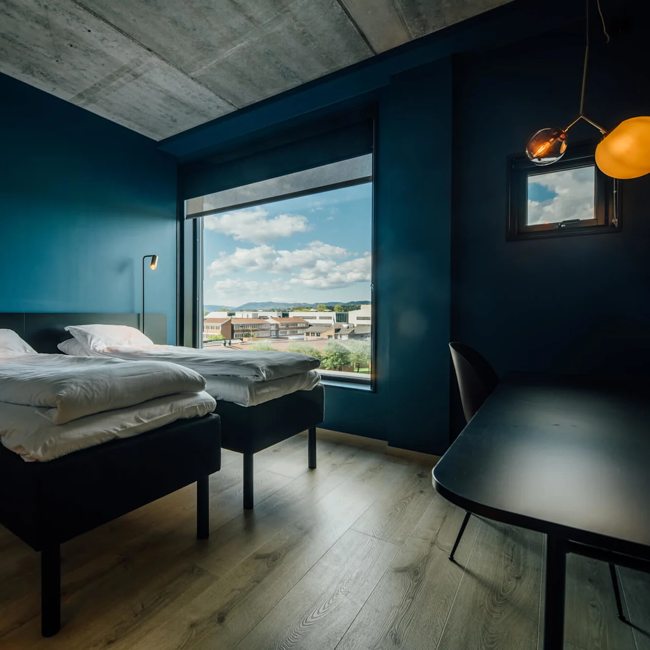 Et moderne hotelværelse med to sirligt redte senge og stilige lamper, med store vinduer der viser et bylandskab under en blå himmel.