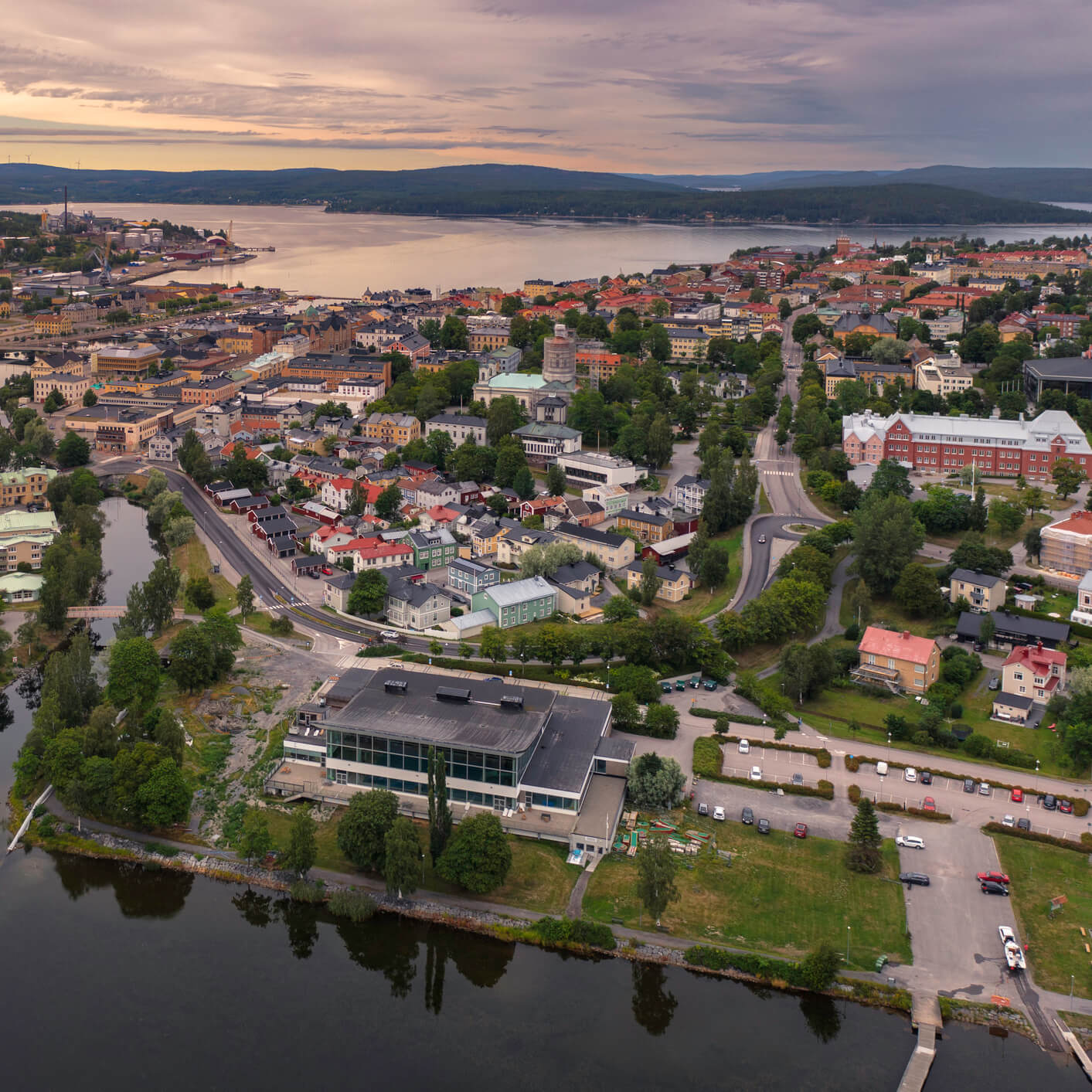 Luftfoto av en by med bygninger, veier, trær og vann i skumringen.