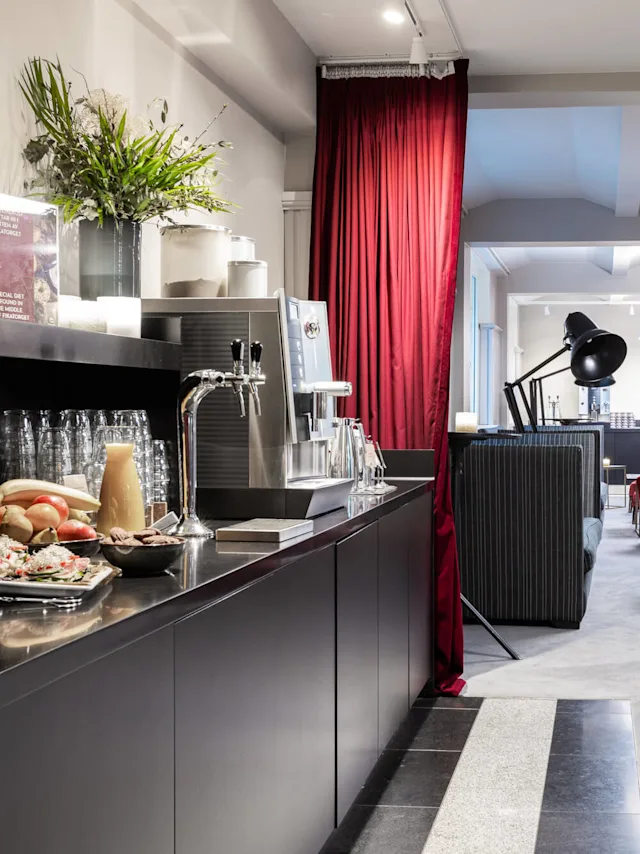 Coffee station with drinks and snacks arranged on a black counter, surrounded by elegant seating with red chairs in a modern, well-lit lounge. Text: "SPECIAL OFFER THE WONDERS OF HOTELNESS".