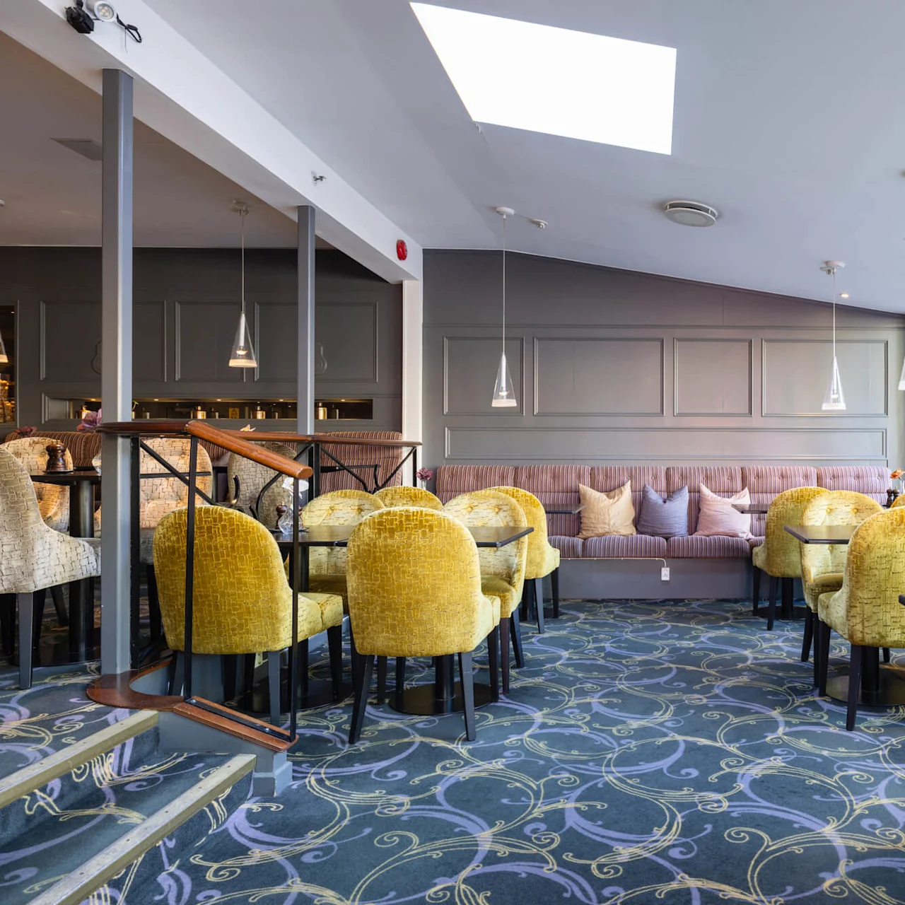 Moderne restaurantinteriør med gule polstrede stoler og mønstret teppe, naturlig lys strømmer inn gjennom store vinduer, pendellamper over.