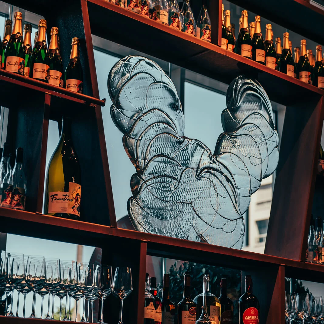 A large wine rack displays multiple bottles of champagne and wine, with an intricate glass sculpture as a centerpiece, set in a dimly-lit bar environment.