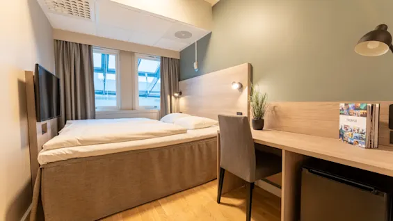 Et moderne hotellrom med en komfortabel seng, et funksjonelt skrivebordsområde med en stol og en TV. En bok om TROMSO ligger på skrivebordet.