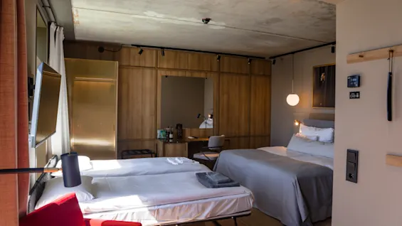 Ett hotellrum har en prydligt bäddad dubbelsäng och en hopfällbar säng. En röd stol och ett skrivbord i trä finns i närheten, med mjuk belysning och modern inredning som skapar en mysig atmosfär.