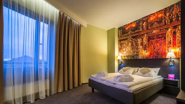 A neatly made hotel bed with decorated pillows and robes sits beneath ornate wall art, flanked by lit sconces. A curtained window offers a view of rooftops.