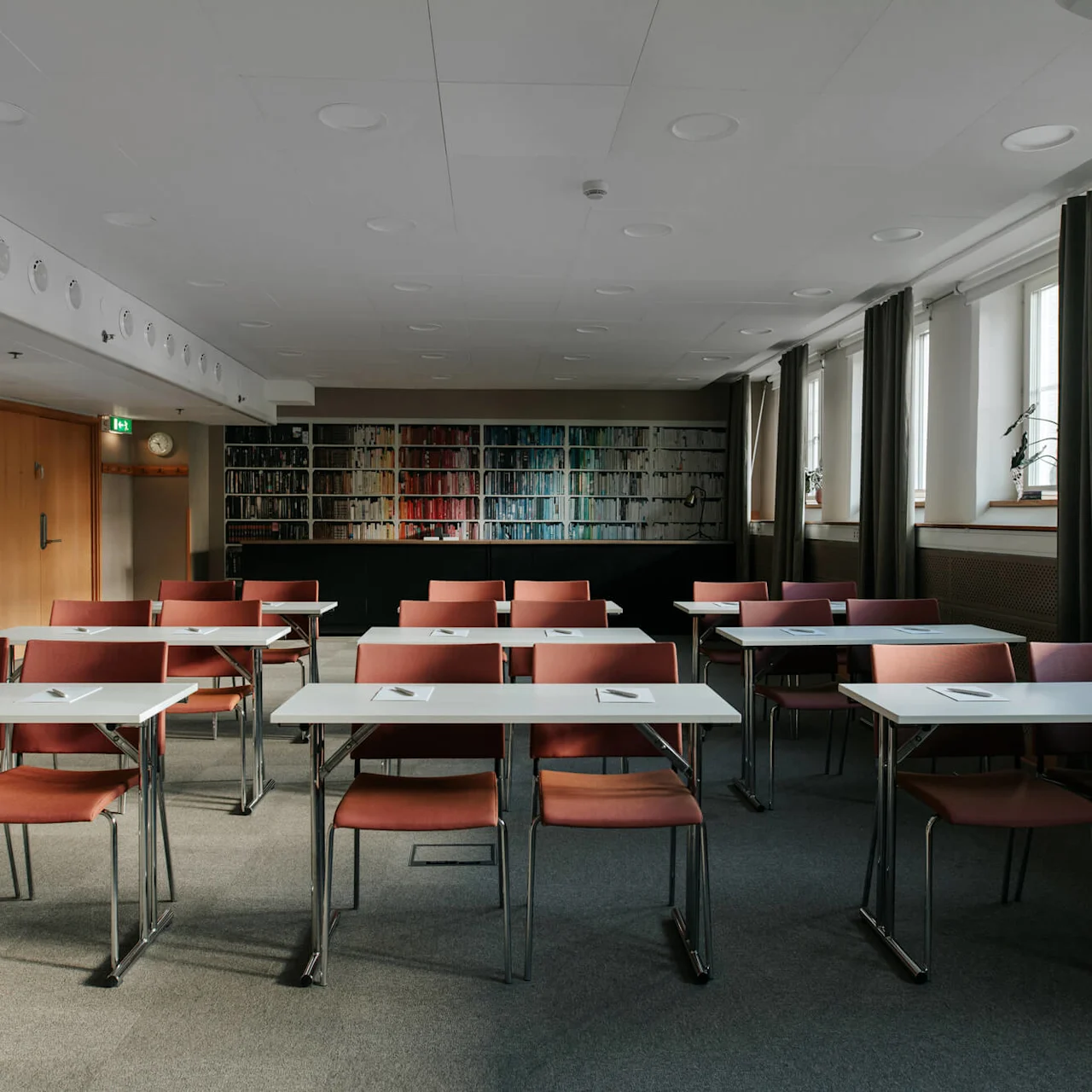 Empty classroom with rows of tables and chairs, facing a bookshelf wall, natural light from windows.