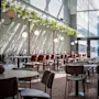 En restaurant med borde og stole pænt arrangeret. Sollys strømmer gennem store vinduer og kaster mønstre på gulvet. Hængende lamper og grønne planter skaber en hyggelig stemning.