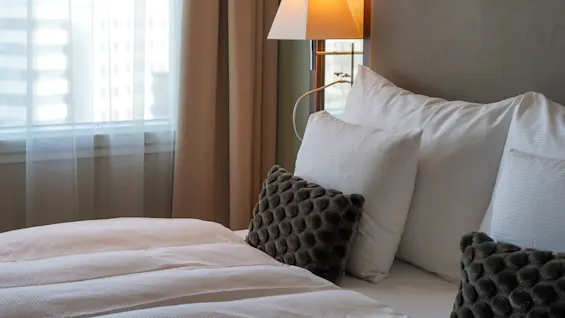 A neatly made bed features white pillows and textured dark cushions, beneath a warm-lit wall lamp. Nearby, sheer curtains cover a sunlit window.