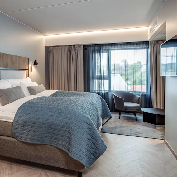 En pent oppredd seng med blått teppe og puter står i et rolig hotellrom, sammen med en stol og bord ved store vinduer med tynne gardiner.