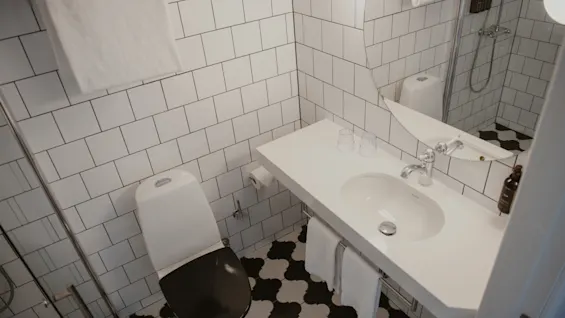 Toalett med svart sits, bredvid en vit vask med en spegel ovanför på en kaklad vägg. Handdukar och glas står på bänken, i ett badrum med rutiga golvplattor.