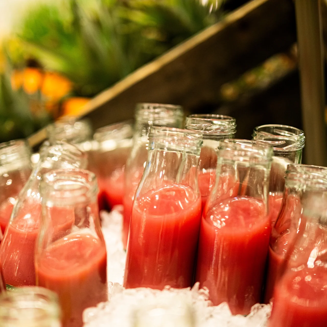 Glass bottles filled with red juice stand upright in a bed of crushed ice, surrounded by greenery and blurred fruits in the background, possibly at a market or buffet.