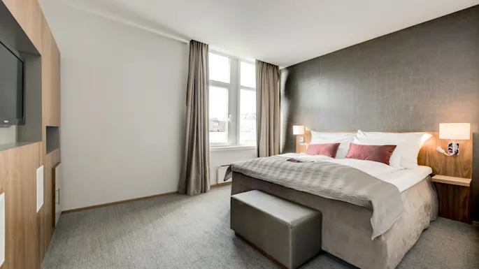 A bed with white linens and red pillows is neatly made against a dark accent wall. Two lamps provide light; curtains frame a window. Modern decor surrounds in a tidy bedroom.