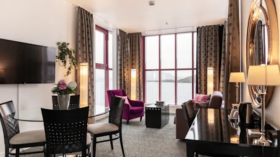 Tilava hotellihuone, jossa on ruokailutila, oleskelutila ja suuri ikkuna, josta on luonnonkaunis näkymä järvelle ja vuorille.