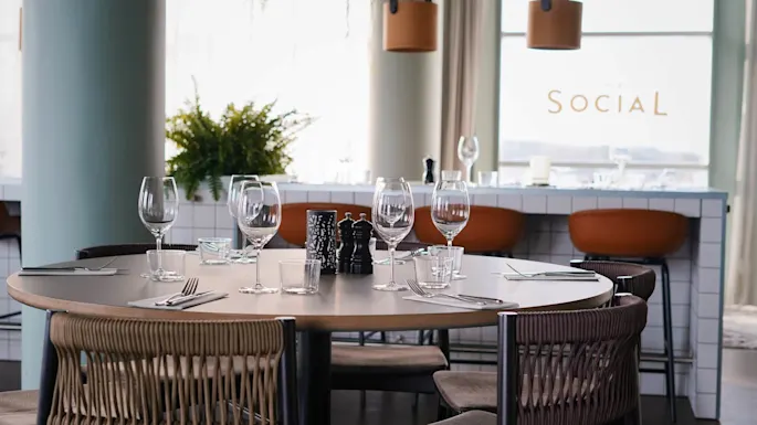 A round dining table set with glassware and cutlery, surrounded by chairs. Background features a bar with orange stools and text reading “SOCIAL” on the window.  