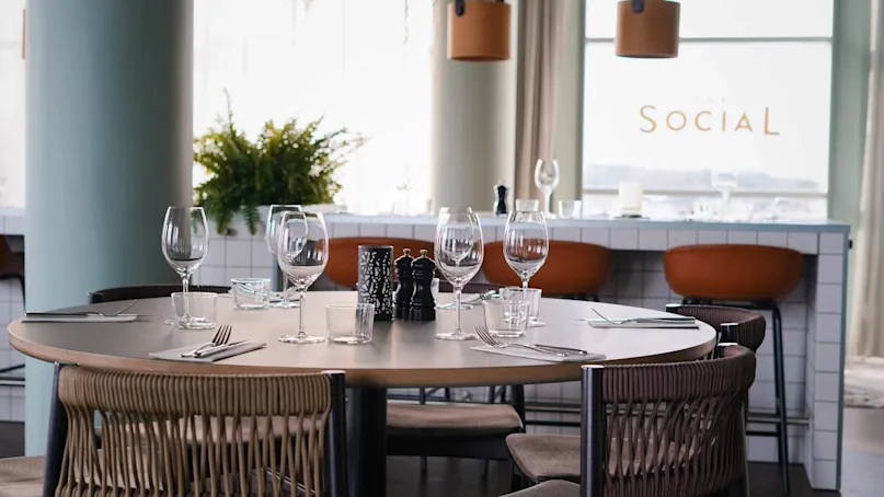 A round dining table set with glassware and cutlery, surrounded by chairs. Background features a bar with orange stools and text reading “SOCIAL” on the window.  