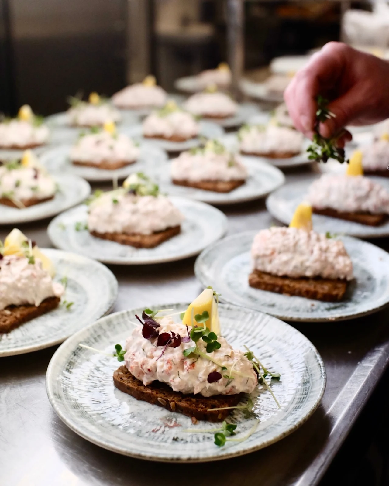 English: Open-faced sandwiches with creamy topping and lemon wedge on plates. Hand garnishing with herbs in a kitchen setting.