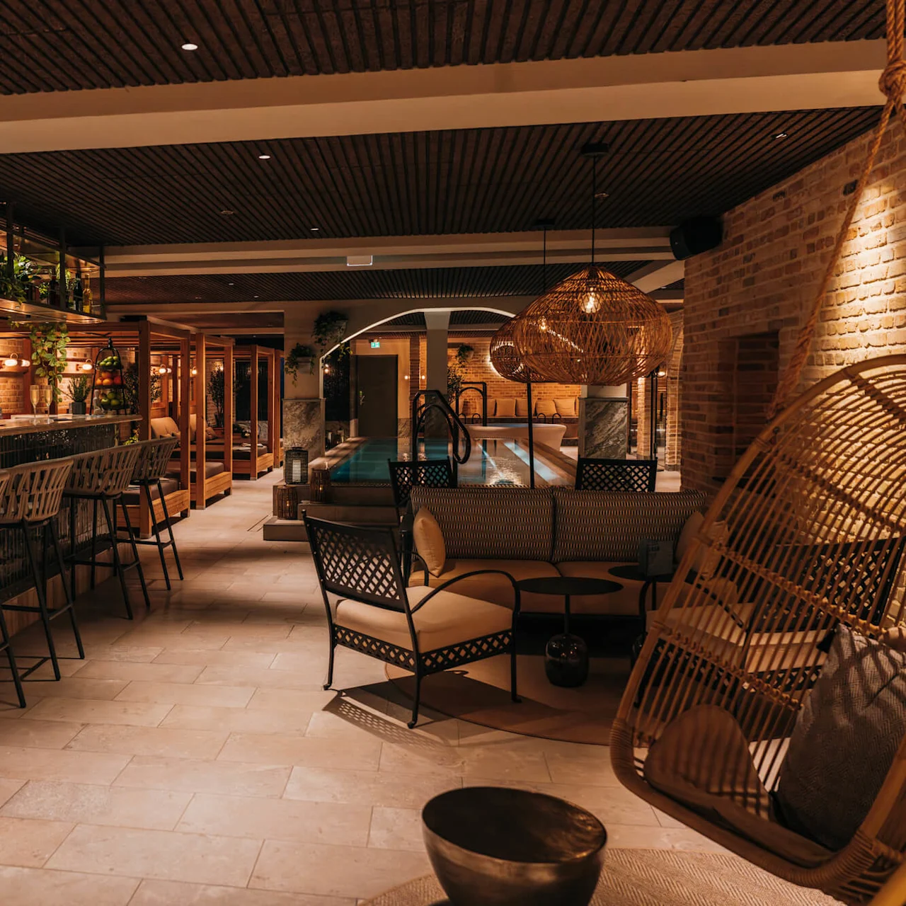 Bar with high stools, backed by shelves with plants and bottles; adjacent to a cozy seating area and illuminated indoor pool in a warmly-lit modern lounge setting. 