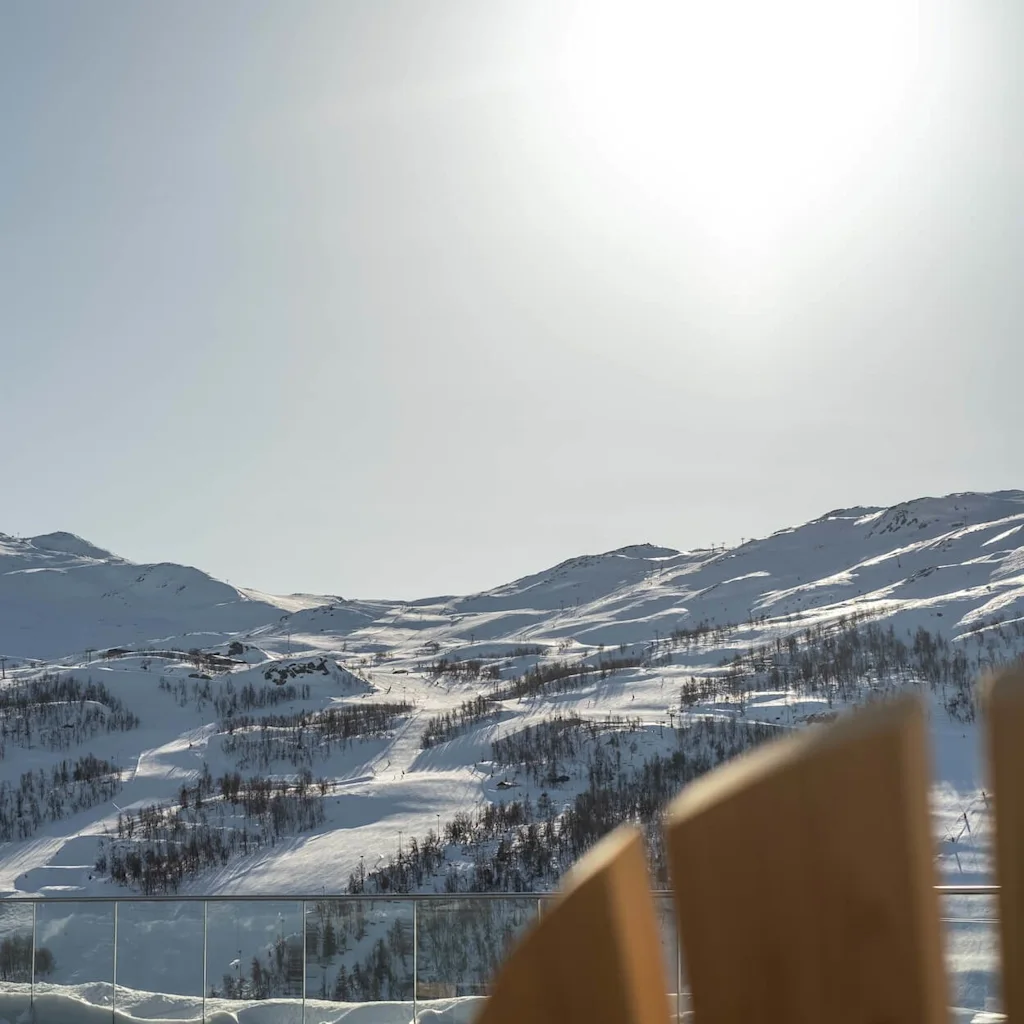 Snötäckta berg står stilla under en klar himmel, med solljus som lyser upp trädkantade sluttningar. Träkonstruktioner syns delvis i förgrunden, vilket antyder en utsiktsplats eller terrass.