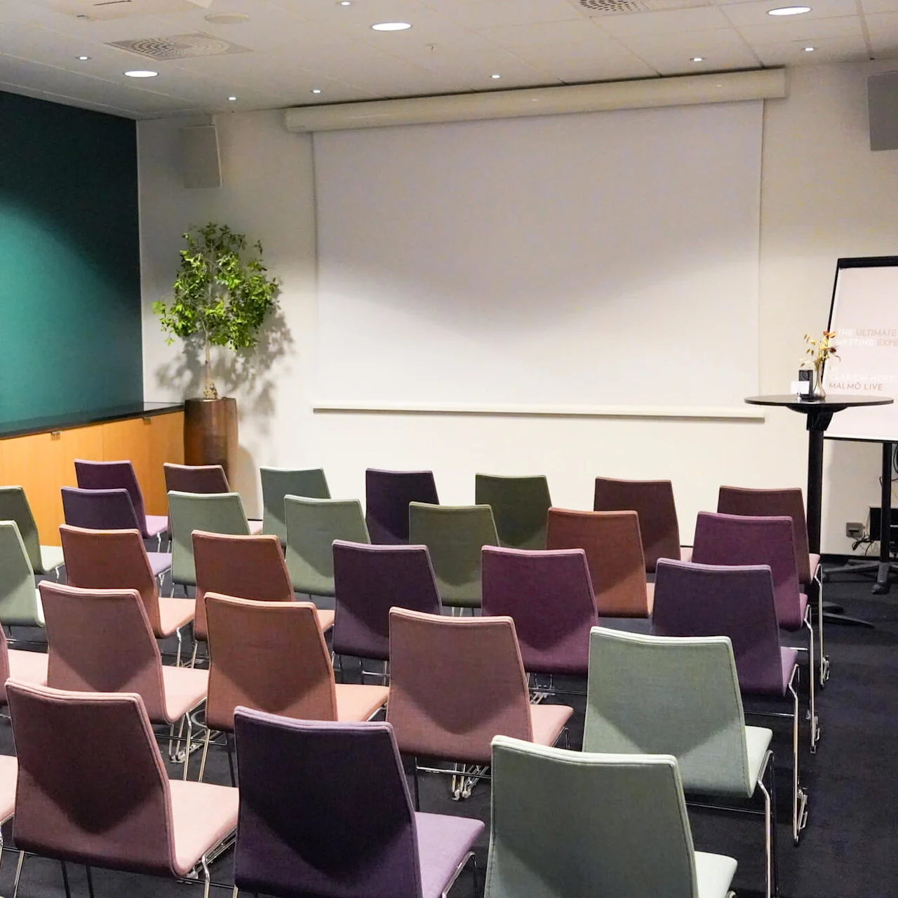 Rader med fargerike stoler vender mot en stor lerret i et konferanserom. Et flippover står det: "THE ULTIMATE MEETING EXPERIENCE" og "MALMO LIVE." En potteplante står i hjørnet.