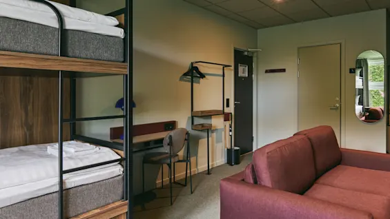 A modern hotel room featuring bunk beds, a desk with a chair, and a comfortable red sofa, designed for relaxation.