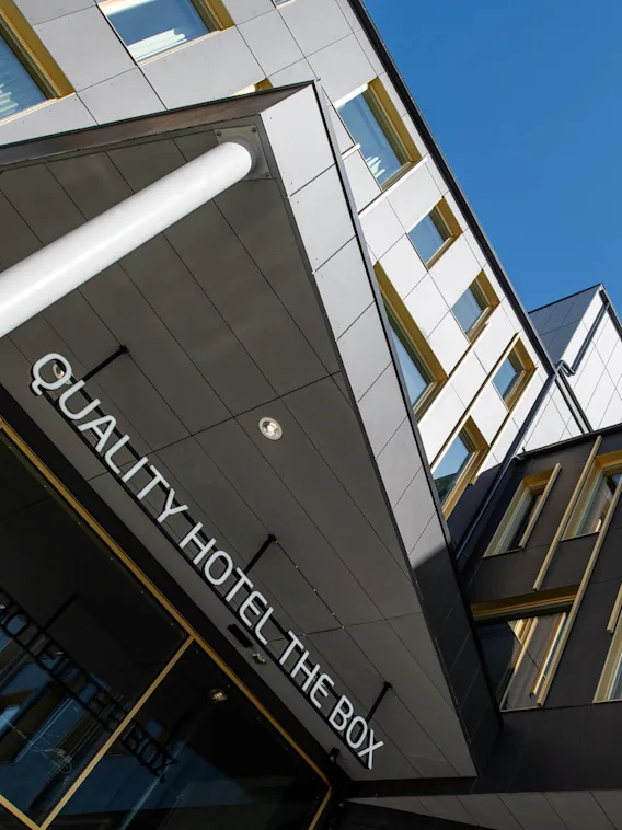Fasaden på en modern byggnad med stora fönster; ingången visar "QUALITY HOTEL THE BOX" i tydliga bokstäver. Himlen är klarblå och framhäver byggnadens geometriska design.