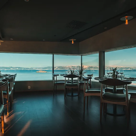 Spisebord er dekket med glass og bestikk, vendt mot store vinduer som gir en naturskjønn utsikt over et rolig hav og fjerne fjell under en klar himmel. Interiøret er svakt opplyst.