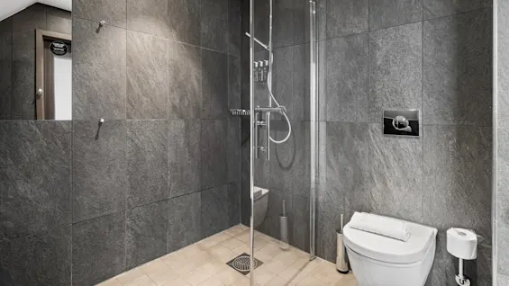 Et moderne badeværelse har en dusjkabin med glass og elegante armaturer. Grå steinfliser dekker veggene, og et hvitt toalett med et håndkle oppå er i nærheten.