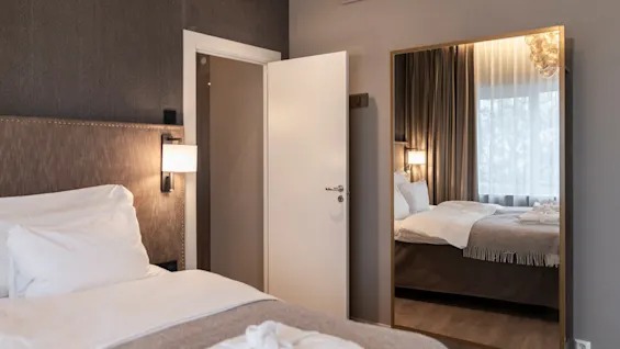 A cozy hotel room at Home Hotel Grand Bodø with a neatly made bed, a bathrobe, and a large mirror reflecting a window.