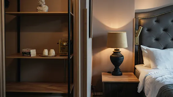 A bedside lamp casts a warm glow beside a tufted headboard and white pillows. Nearby, a shelf holds a bust, books, stacked cups, and a gold coffee machine.