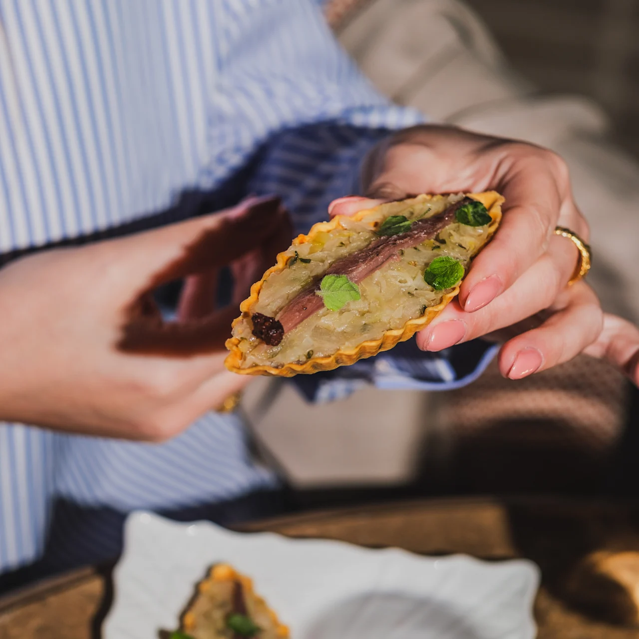 A person holds a small tart topped with anchovies and herbs, wearing a striped blue shirt. Another tart sits on a white plate below on a table.