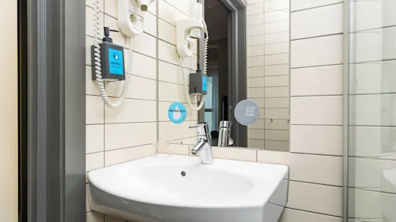 A white sink with a modern faucet is mounted on a tiled bathroom wall. Above, a mirror shows two stickers: “DRINK MORE WATER” and “GO EASY ON THE PLANET.” A soap dispenser and hair dryer are adjacent.