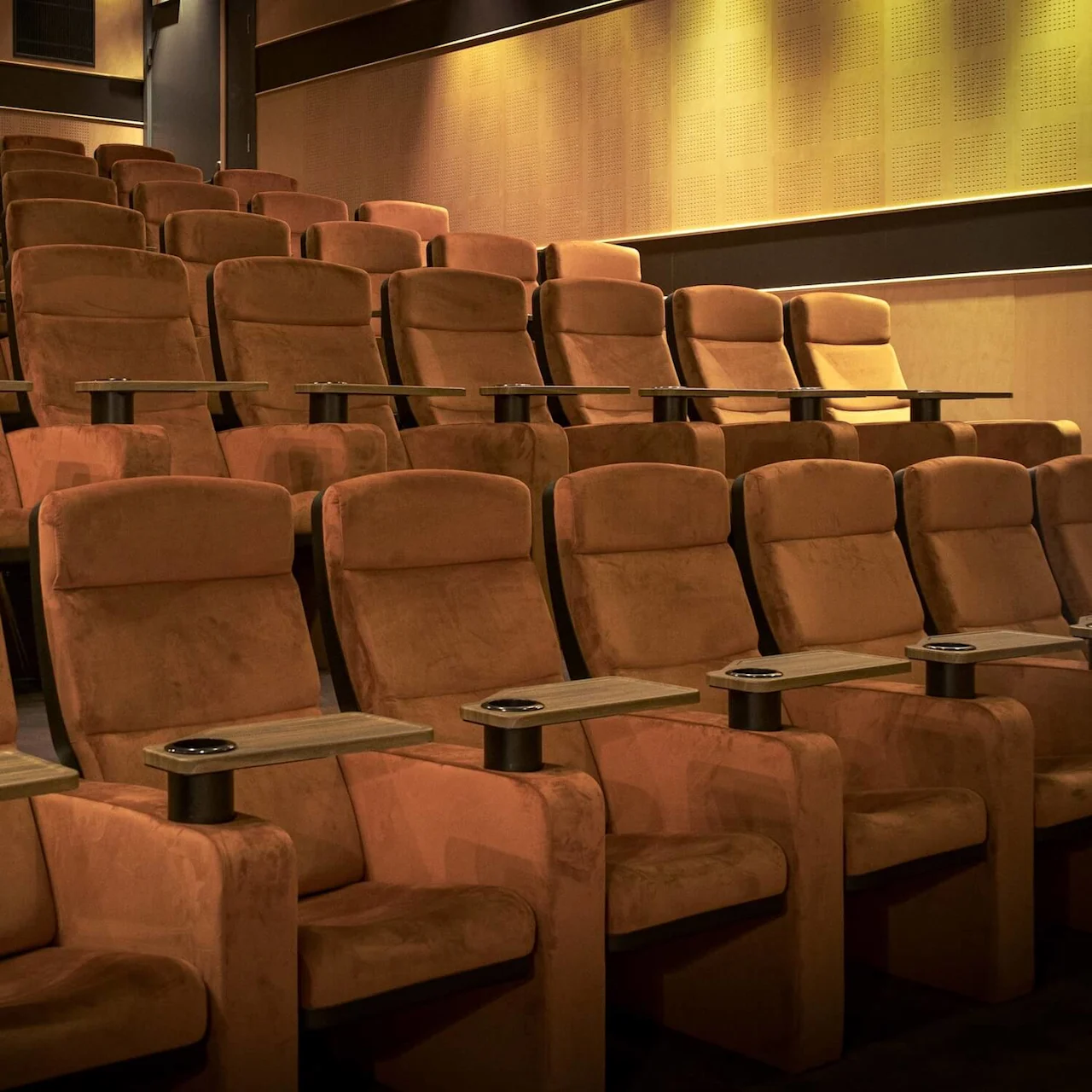 Plush cinema seats with wooden armrests and cup holders are arranged in rows within a dimly lit theater, creating an inviting atmosphere for movie-goers.