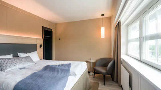 A bed with a gray blanket and pillows is set in a minimalist hotel room. A chair and small table are near a large window with curtains.